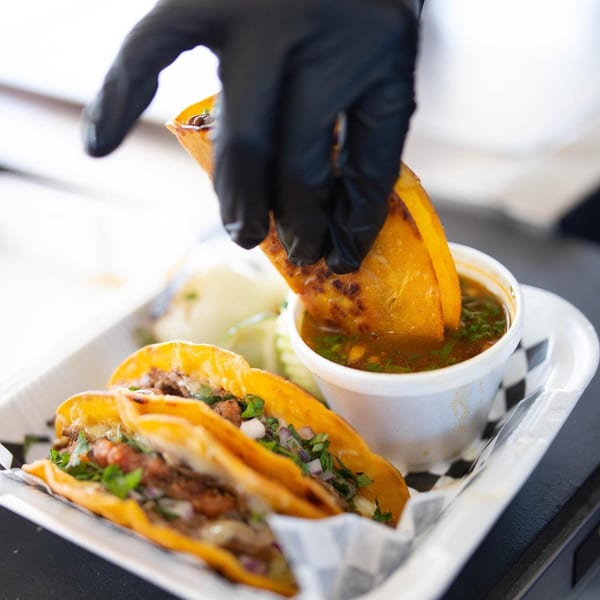 A gloved hand dips a Tacos Chapala birria taco into a small cup of consommé.