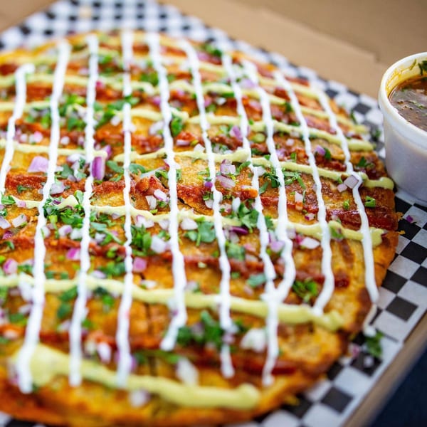 A large Mexican pizza with red sauce, white and green drizzles, cilantro, and red onions, with a side of broth.
